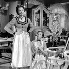 Zwei Frauen in traditionellen Kleidern. Eine Frau steht, die andere kniet und hält den Rock der Stehenden. Ein ländlicher Raum.