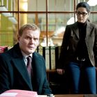 Zwei Personen in einem Büro. Eine sitzt an einem Tisch mit Akten, die andere steht daneben und trägt eine Brille.