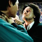 Zwei Männer stehen sich nah gegenüber; einer hält den anderen am Hals, beide wirken angespannt.