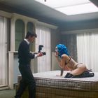 Ein Mann fotografiert eine Frau mit blauen Haaren auf einer Matratze in einem Raum mit großen, weißen Vorhängen.