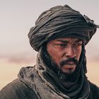 Es gibt keinen Text zu erwähnen. Ein Mann mit Turban schaut ernst, im Hintergrund eine Wüstenlandschaft.