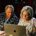 Zwei ältere Personen schauen mit ernster Miene auf einen Laptop mit Apfel-Logo, umgeben von bunten Stiften und einer Schere.