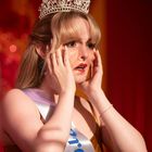 Eine Frau mit Krone und Schärpe schaut überrascht oder besorgt und hält ihre Hände an ihr Gesicht.