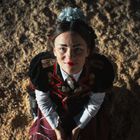 Eine Person in traditioneller Kleidung schaut lächelnd nach oben, mit einem dekorativen Kopfschmuck.