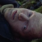 Ein Mann liegt mit offenem Mund und weiten Augen auf moosigem Boden, trägt einen dunklen Mantel und Rollkragenpullover.