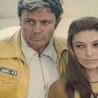 Text "5005-АТ" auf gelber Jacke. Zwei Personen, ein Mann in gelber Jacke, eine Frau daneben, beide schauen ernst.