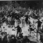 Kein Text zu erkennen. Eine Boxszene mit vielen Menschen im Ring und um den Ring herum. Viel Gedränge und Aufregung.