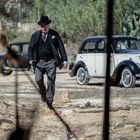 Ein Mann in eleganter, altmodischer Kleidung und Hut geht über einen staubigen Boden. Im Hintergrund sind zwei Oldtimer zu sehen.