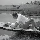 Ein Mann und eine Frau liegen eng beieinander auf einem Steg an einem ruhigen See, umgeben von hügeliger Landschaft.