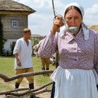 Eine Frau mit Kopftuch und Blumenkleid trinkt aus einer Kelle. Ein Mann in traditioneller Kleidung im Hintergrund.