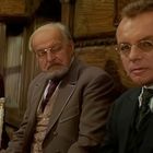 Fünf Männer in älterer Kleidung sitzen in einem Holzraum, drei schauen nachdenklich, einer hält einen Stock.