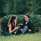 Zwei Personen sitzen auf einer Wiese vor einer efeubewachsenen Mauer, eine liest aus einem Buch vor.