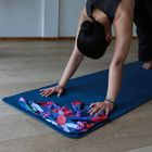 Frau macht Yoga auf blauer Matte, trägt dunklen Anzug, bunter Schal liegt vorne. Hände und Füße stabil am Boden.