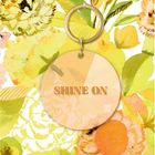 „SHINE ON“ steht auf einem runden Anhänger. Buntes, blumiges Hintergrundmuster mit leuchtenden Farben.