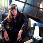 Ich kann keine Texte im Bild erkennen. Eine Person mit Fliegermütze und Brille sitzt lässig auf einem Sessel in einer Scheune.