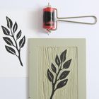 Eine schwarze Pflanzenillustration auf weißem Papier, daneben eine Druckplatte mit Farbroller.