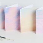 „Liebe Maya“ steht auf einer aufgeklappten Karte. Links davon ein Stift, im Hintergrund Karten in Pastelltönen.