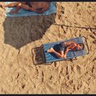 Ein Junge liegt auf einer blauen Liege im Sand, daneben entspannen andere Personen unter einem Sonnenschirm.