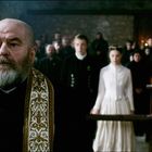 Ein Priester im Vordergrund mit verzierter Stola, Menschen in schwarzer Kleidung und brennende Kerzen im Hintergrund.