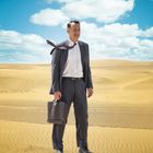 Ein Mann in Anzug steht in einer Wüste. Krawatte weht im Wind, Aktentasche in der Hand, blauer Himmel mit Wolken.