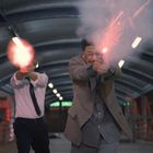 Ich kann keinen Text erkennen. Zwei Männer in Anzügen und zwei weitere Personen schießen in einem überdachten Gang.