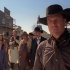 Eine Gruppe von Menschen in Westernkleidung steht auf einer staubigen Straße einer Westernstadt.