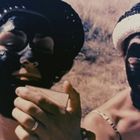 Zwei Personen tragen schwarze Gesichtsmasken und Strohhüte und halten kleine Spiegel in der Hand. Hintergrund: gelber Wiesenboden.