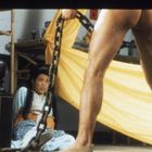 Eine sitzende Frau schaut ängstlich zu einem muskulösen Mann mit Ketten. Hintergrund mit gelbem Tuch und Holzgerüst.