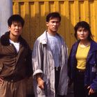 Drei Personen stehen vor einer gelben Metallwand. Die mittlere Person hält eine Waffe. Alle blicken ernst in die Ferne.