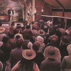 Mehrere Menschen in Hüten und historischer Kleidung versammeln sich, während ein Mann auf einer Bühne spricht.