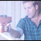 Eine Person zielt mit einem rot-schwarzen Gerät, Funken sprühen. Sie trägt ein grün kariertes Hemd, Hintergrund aus Holzwänden.