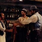 Das Bild zeigt drei Männer in einem Saloon. Im Hintergrund stehen Spirituosenflaschen. Einer der Männer hält einen Sattel.