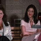 Zwei Frauen sitzen nebeneinander auf Holzbänken und lächeln. Sie tragen elegante, helle Kleidung und befinden sich in einem Innenraum.