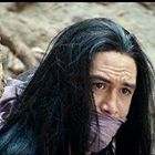 Langer, dunkler Haarträger mit einem lila Schal über Mund und Nase, zwischen Bäumen und Felsen blickend.