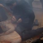 Menschen in einer dramatischen Szene, ein Mann liegt am Boden und wird von einem anderen gehalten. Düstere, verschwommene Umgebung.