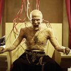 Ein furchteinflößender, ausgemergelter Mann sitzt auf einem Thron, verbunden mit roten Kabeln, in einer unheilvollen Umgebung.