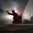Ein Mann in roter Arbeitskleidung kriecht in einem engen Schacht, hält ein leuchtendes grünes Licht. Er trägt einen Schutzhelm.