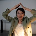 Es sind keine Texte vorhanden. Eine Frau in einem grünen Blazer formt ein Dreieck mit den Händen über dem Kopf.