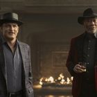 Zwei Männer in Hüten, einer im karierten Anzug, der andere in roter Jacke, stehen vor einem Kamin. Der rechte hält ein Getränk.