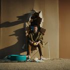 Eine Person sitzt in einem Loch in der Wand, eingehüllt in einen Umhang, mit einem Stock und einem blauen Behälter davor.