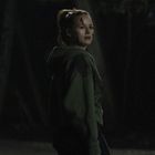 Eine Frau in einem dunklen Wald, trägt einen grünen Hoodie und schaut ernst. Es ist Nacht und die Szene ist düster.