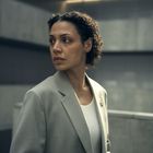 Keine sichtbaren Texte. Eine Frau mit lockigem Haar in einem hellen Anzug steht vor einer modernen, grauen Betonwand.