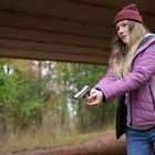 Keine sichtbaren Texte. Eine Frau, in lila Jacke und Mütze, hält eine Pistole in einer bewaldeten Umgebung.