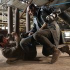 Zwei Männer kämpfen am Boden in einer industriellen Umgebung mit Rohren und Metallstrukturen.