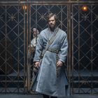 Ein Mann in einem langen, grauen Mantel steht in einem Käfigbereich mit Metallgittern. Eine weitere Person ist im Hintergrund.