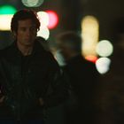 Keine Texte sichtbar. Eine Person in einer Lederjacke steht nachts auf einer belebten Straße mit unscharfen Lichtern im Hintergrund.