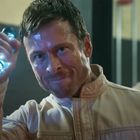 Ein entschlossener Mann hält ein leuchtendes, futuristisches Instrument hoch. Im Hintergrund ist eine weitere Person unscharf.