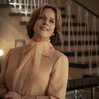 Eine elegant gekleidete Frau lächelt, steht vor einer Treppe mit Barriere und goldenen Details.