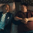 Zwei Männer liegen nebeneinander auf einem Bett mit gemusterten Decken, umgeben von weichem Licht.