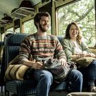 Ein Mann und eine Frau lachen auf einem Sitz in einem Zugabteil, umgeben von Reisetaschen.